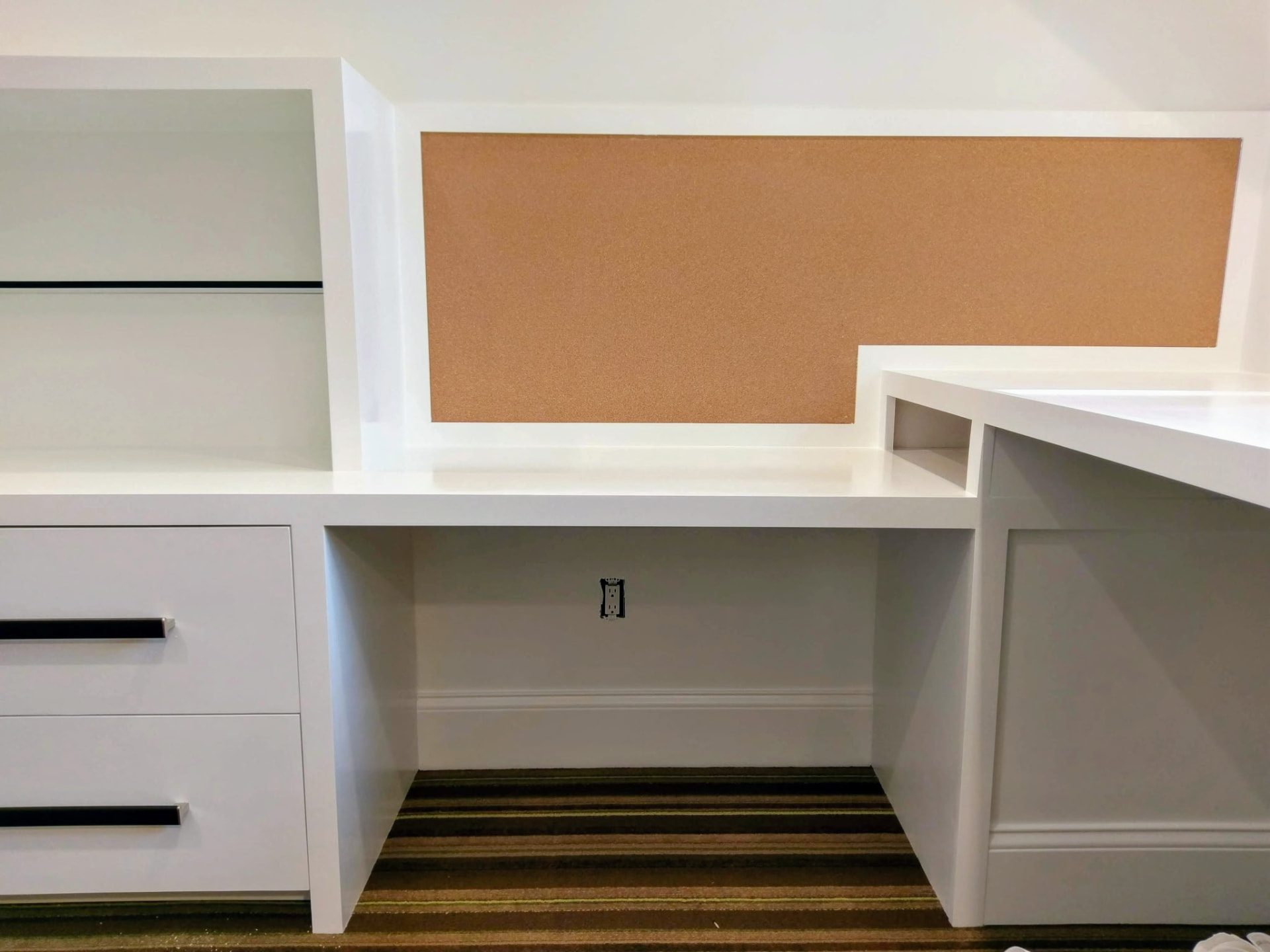 Integrated desk with framed corkboard pin panel, drawer storage and glass display shelving in the white home office built-in cabinetry in Ridgefield, CT