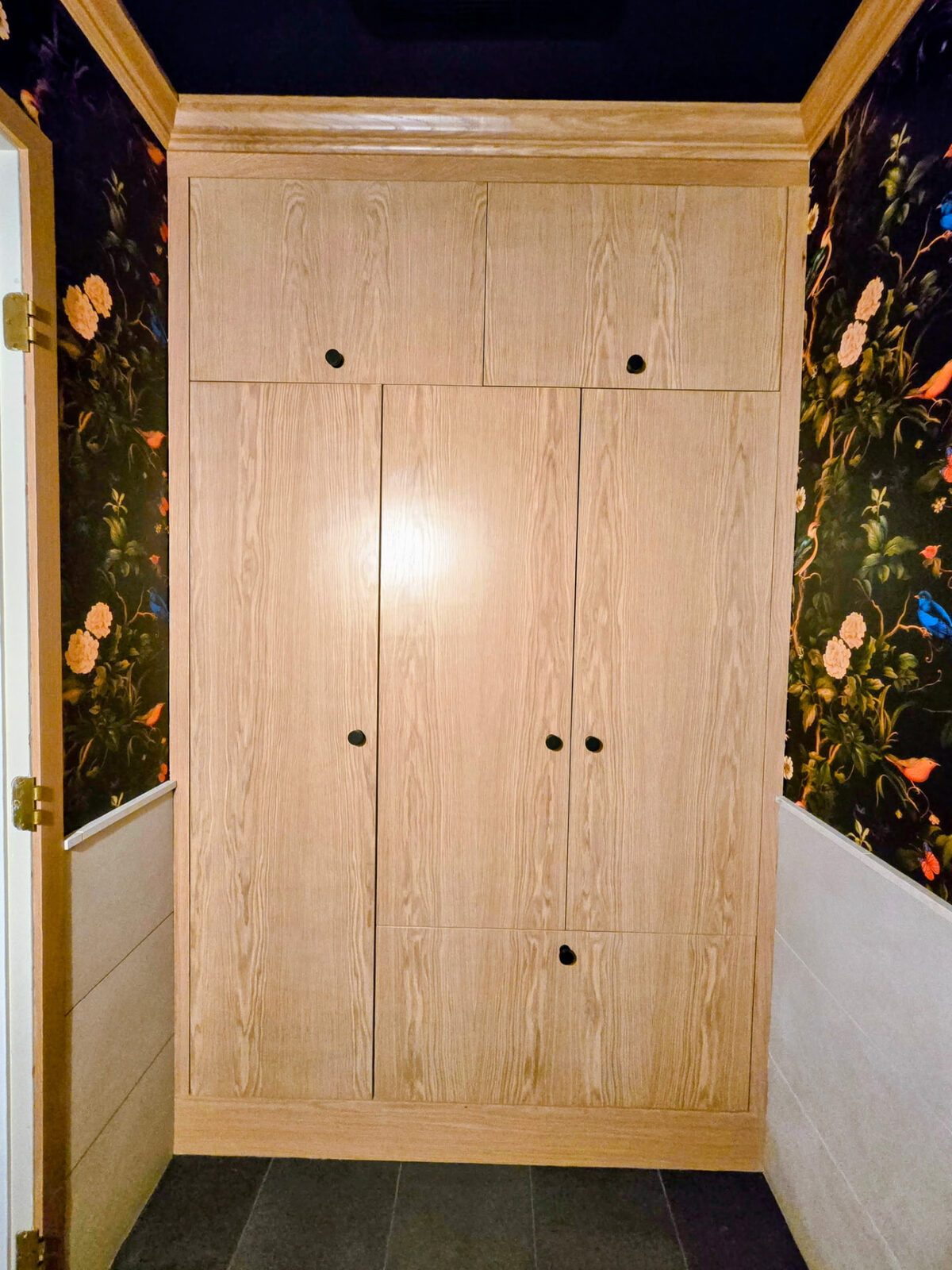 Completed floor-to-ceiling custom white oak closet with crown molding and black knobs, framed by floral wallpaper